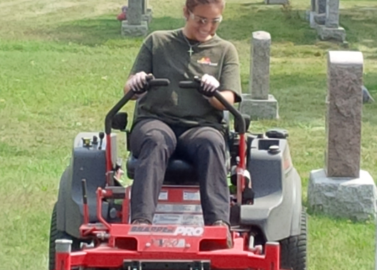 smiling woman riding red lawn mower in cemetery