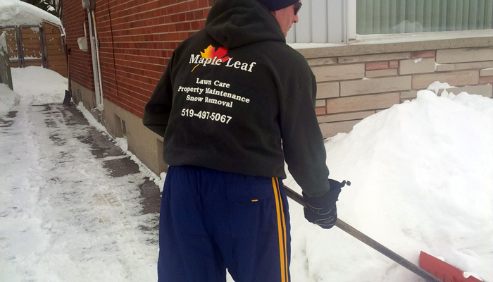 maple leaf lawn maintenance employee in branded black sweater shoveling snow at residential property
